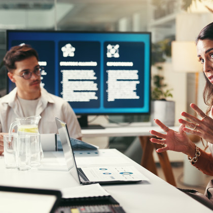 Woman gestures while presenting in a meeting room, with a colleague and a large screen displaying information in the background.