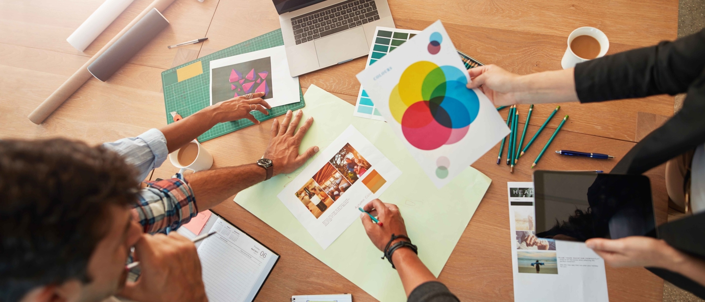 Overhead view of a team reviewing design mockups and colour palettes on a table with a laptop, pencils, and coffee mugs.