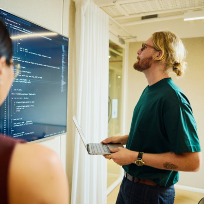 Developer holds a laptop while reviewing code on a large wall-mounted screen during a team discussion in a meeting room.