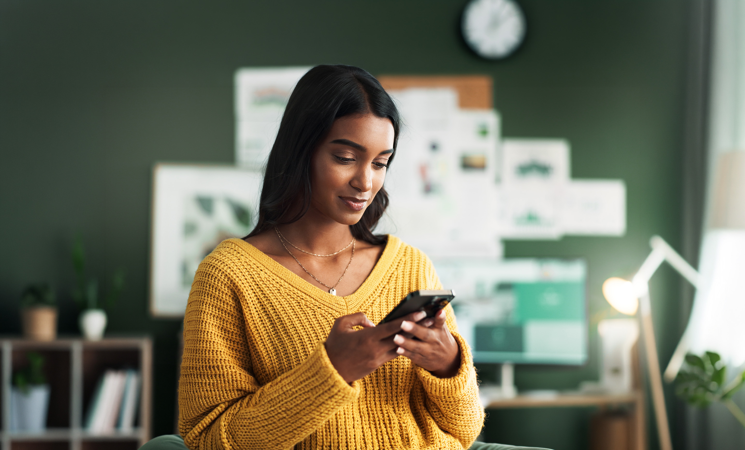 Woman in a mustard-yellow sweater smiles while using a smartphone in a modern office with design boards in the background.