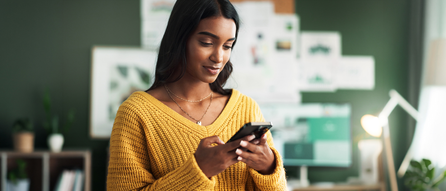 Woman in a mustard-yellow sweater smiles while using a smartphone in a modern office with design boards in the background.